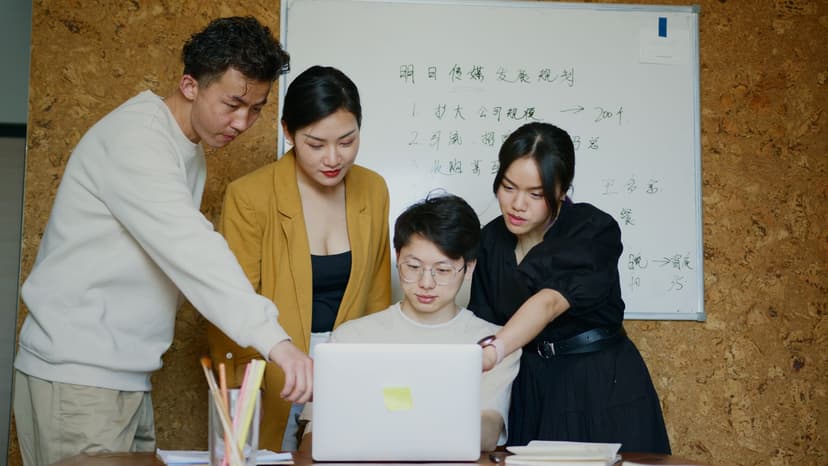 Di open office, seorang developer mengetik cepat sendirian, sementara di belakangnya rekan-rekan tim berkumpul di depan whiteboard mendiskusikan diagram yang rumit