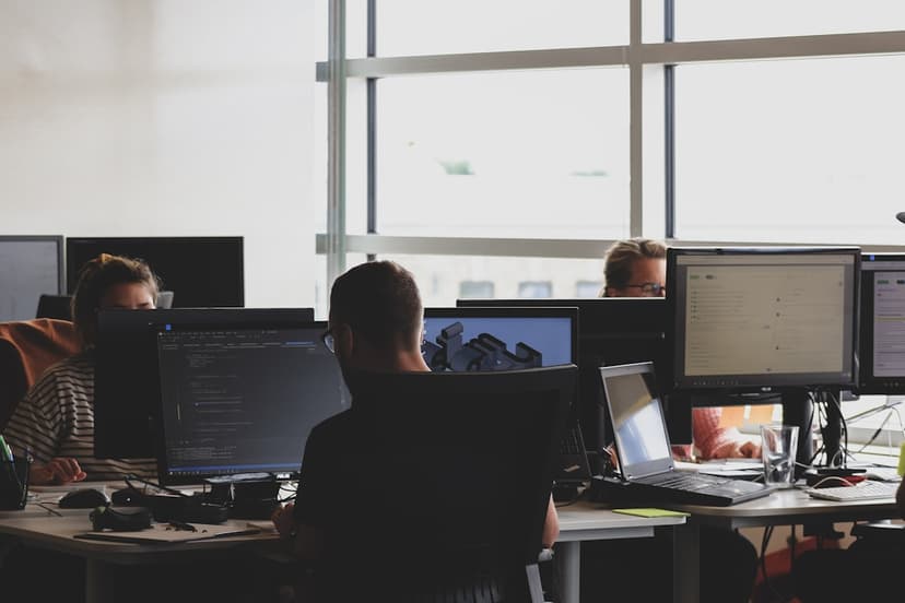 Dua developer di sudut kantor sempit sedang melihat monitor bersama untuk code review, salah satunya menunjuk layar dengan ekspresi tegang